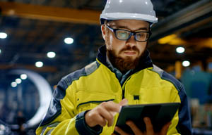 Man wearing a high visibility jacket and a hard hat looking at a screen in a warehouse.